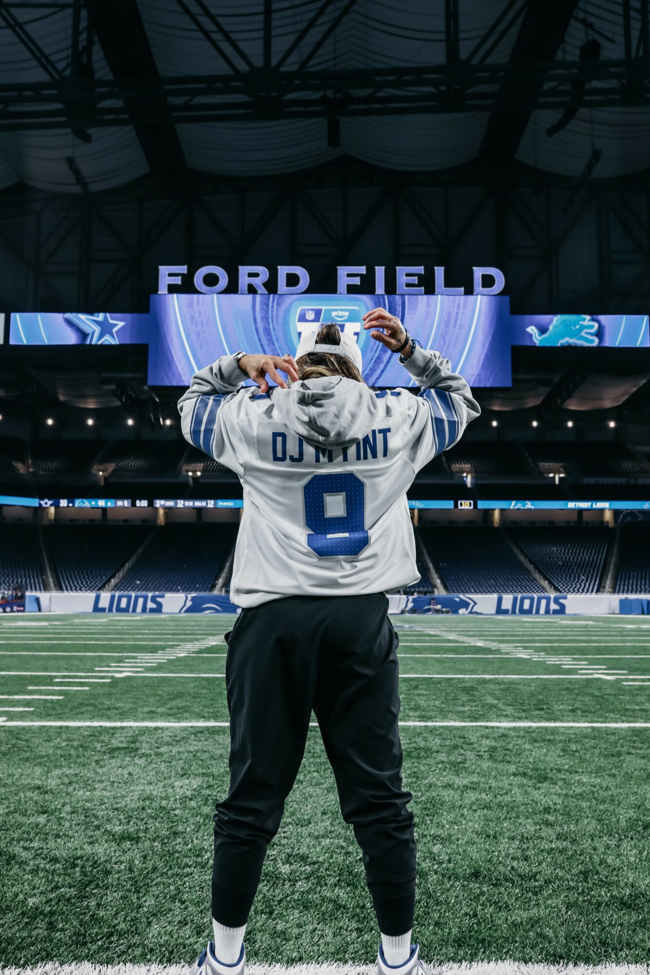 Person in a Detroit Lions jersey holding a helmet on a football field at Ford Field, with stadium and scoreboard in background.