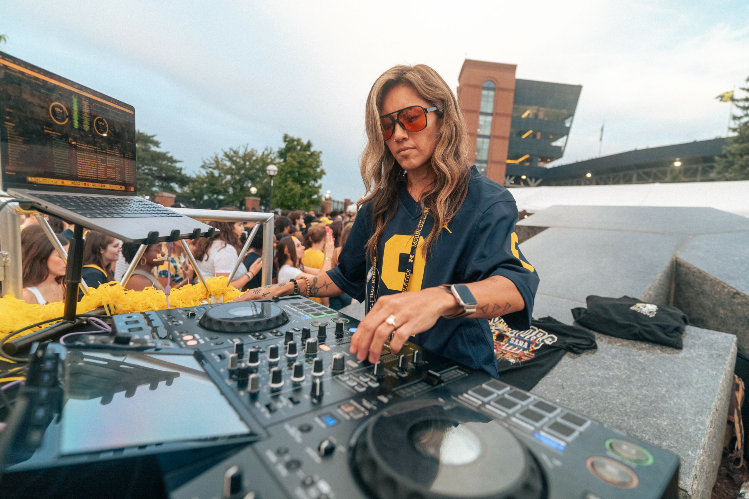 Woman DJ wearing sunglasses and a jacket, operating DJ equipment outdoors during daytime, with a crowd and modern building in background.
