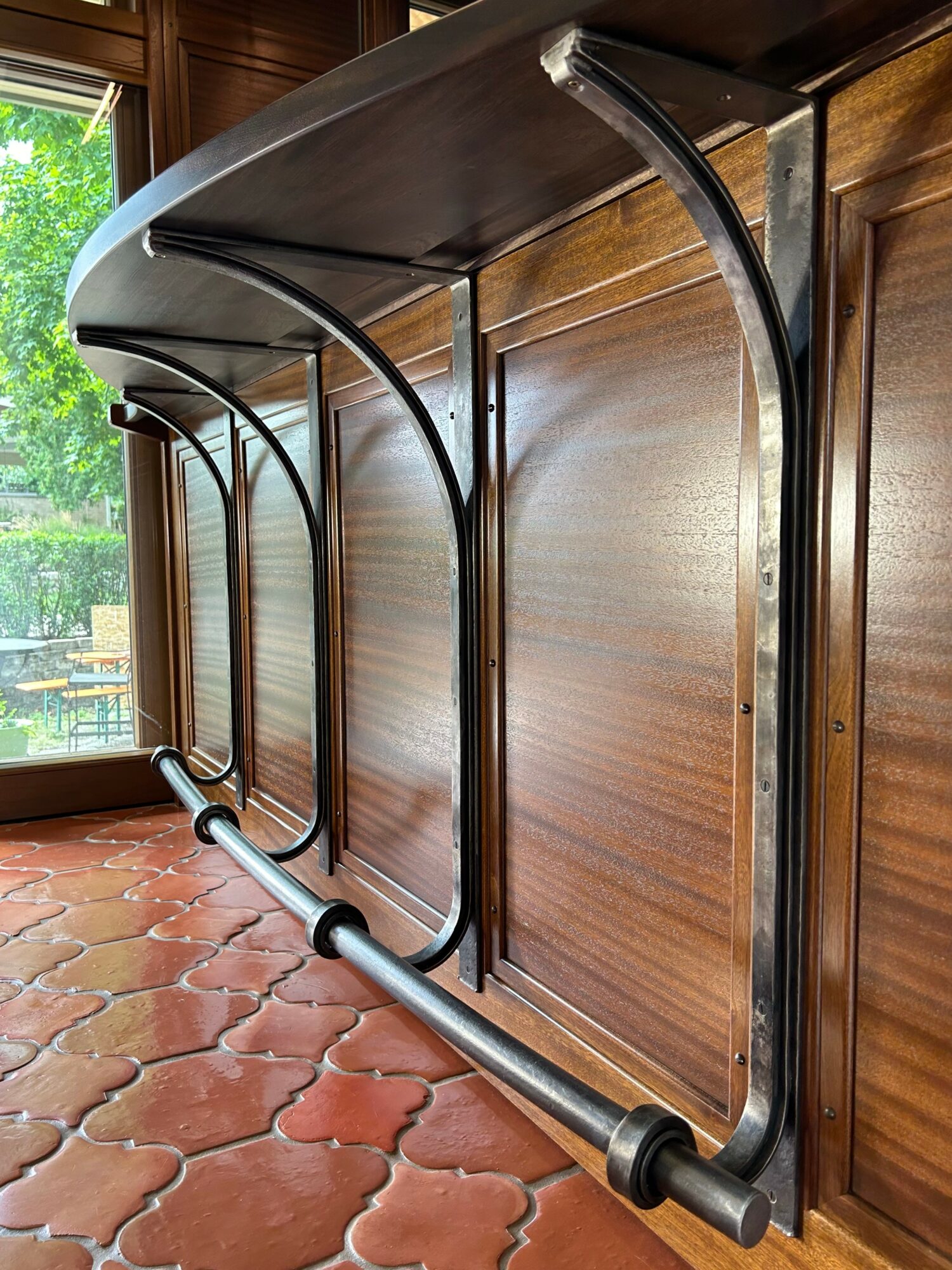 Metal and glass table with curved legs on a wooden floor near a window with greenery outside.