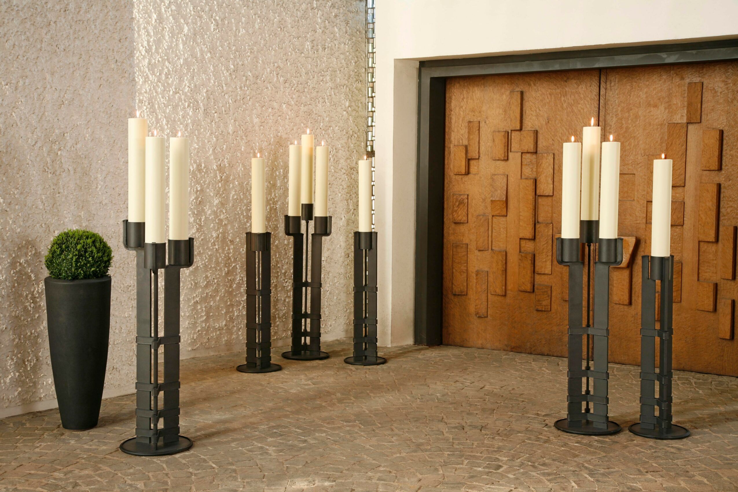 Six black candle holders with white candles arranged near a wooden door and a potted plant, on a concrete floor.