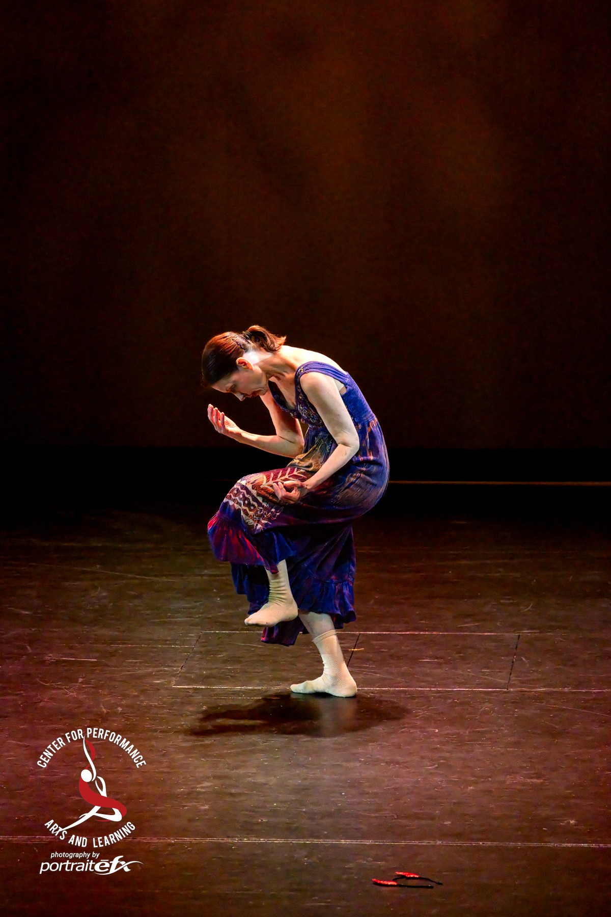 A dancer in a blue and patterned costume performs on stage with a dark background, balancing on one foot with arms raised.