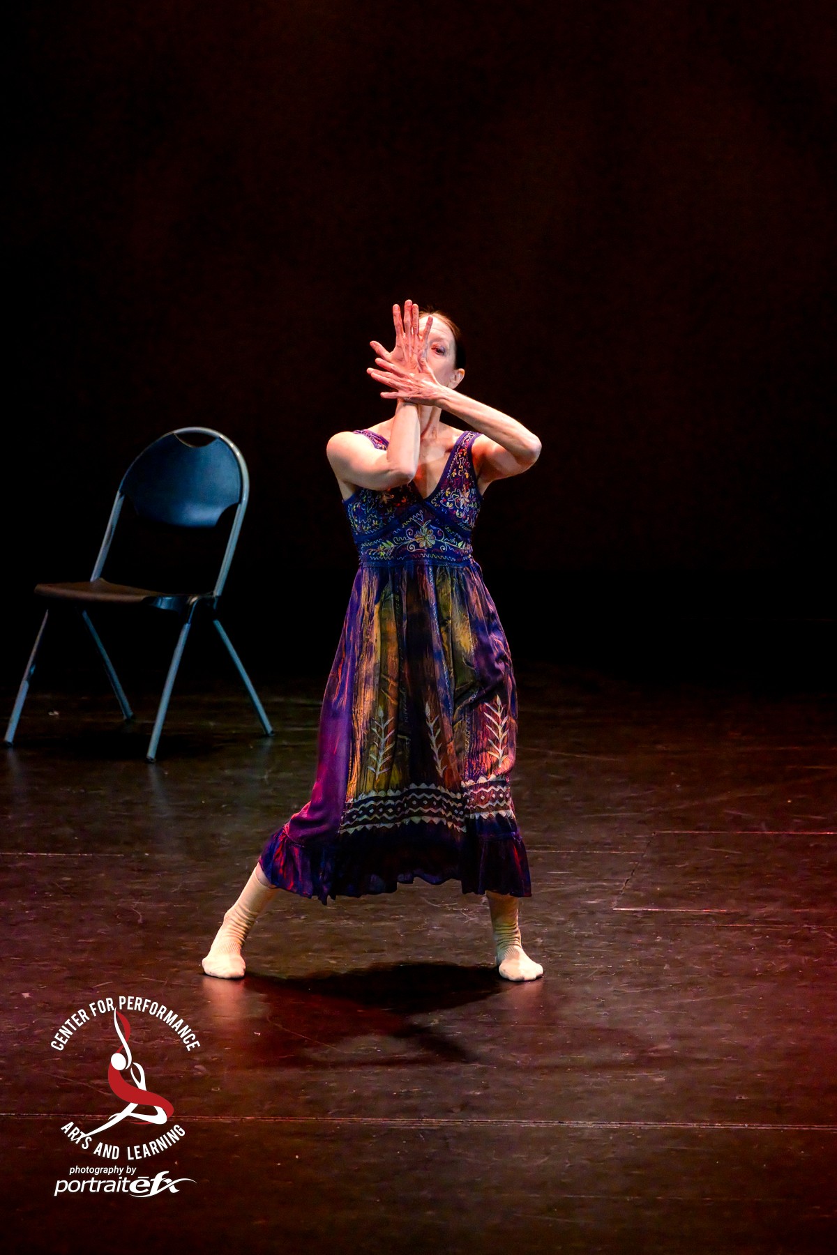 Person dancing on stage with a chair nearby, wearing a colorful dress, hands covering face, dark background.