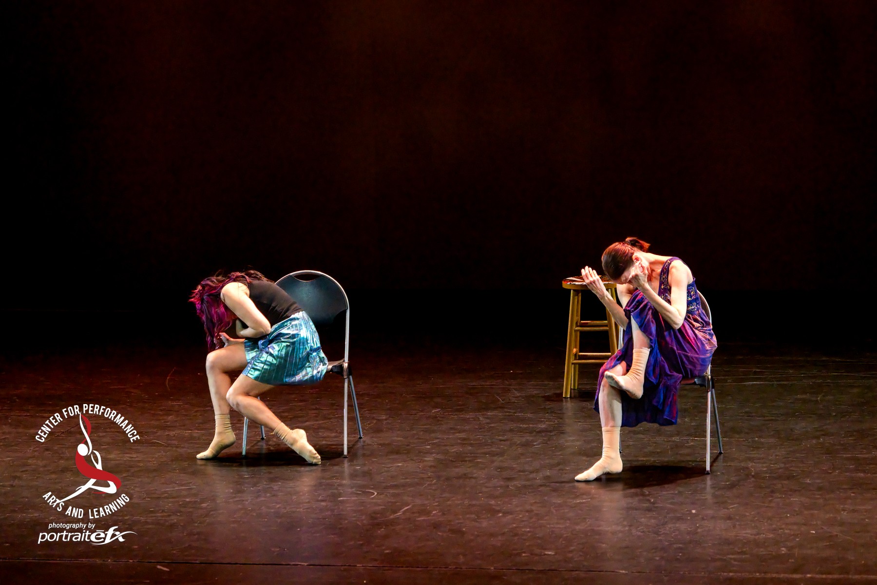 Two performers sit on chairs on stage, leaning forward with heads down and hands on their heads, in a dance performance.