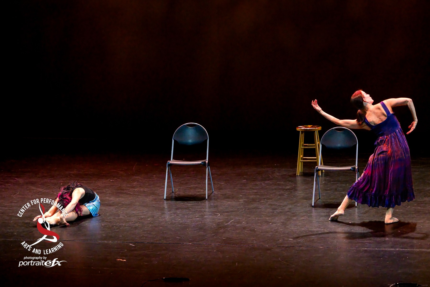 Two dancers perform on stage with chairs and a stool, one lying on the floor and the other standing with an arm raised.