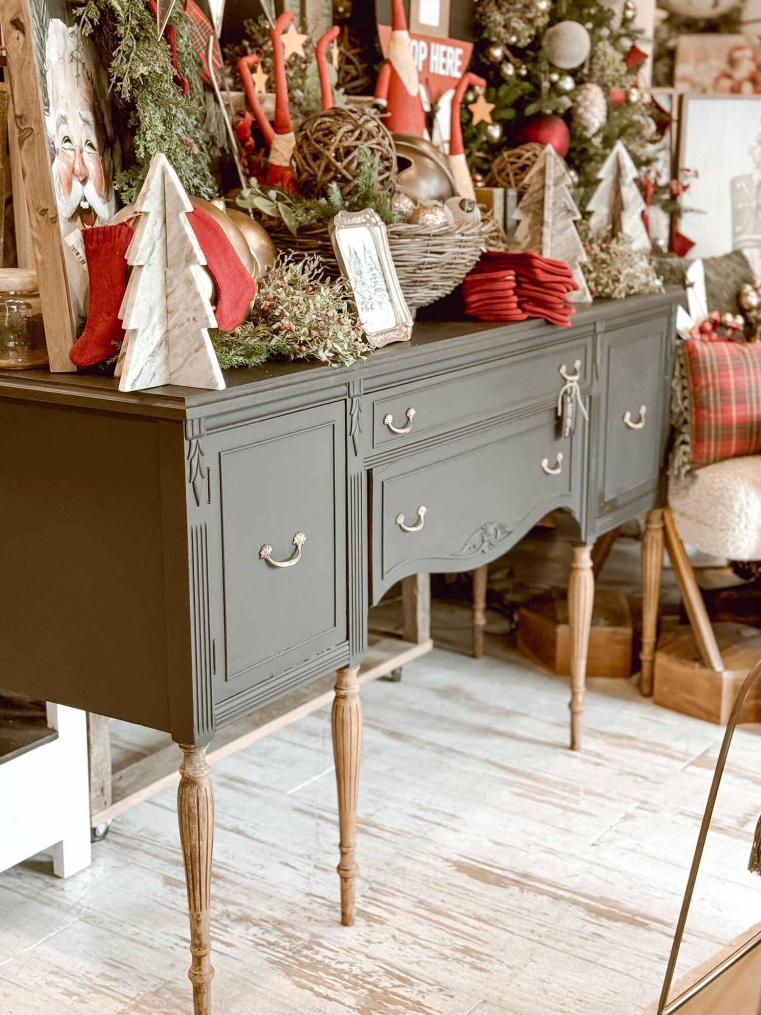 Decorated sideboard with Christmas ornaments, trees, and red accents, in a festive room setting, with a chair nearby.