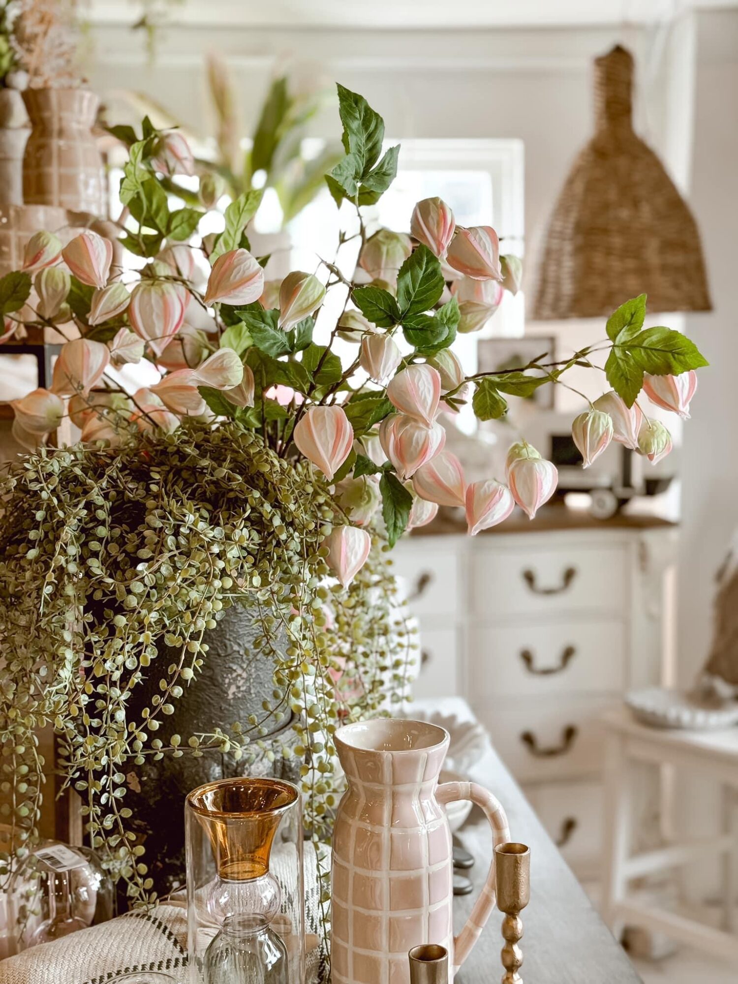 Vase with pink and white flowers and green leaves on a table, with a kitchen in the background.