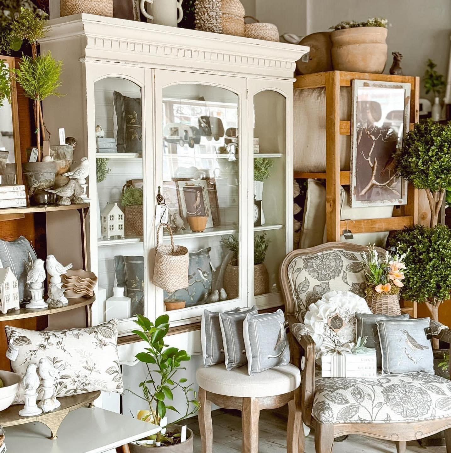 Display cabinet with glass doors, surrounded by furniture and plants in a cozy room, decorated with flowers and ornaments.