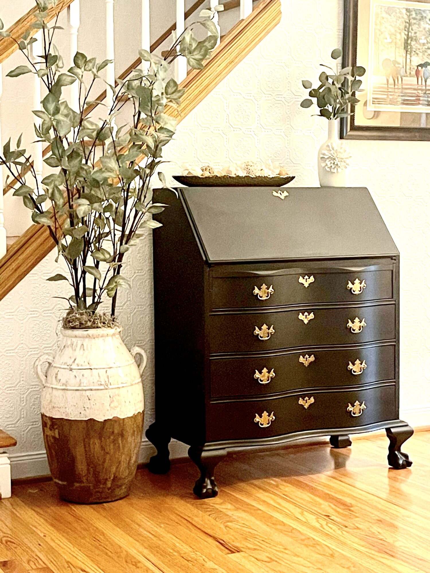 Black dresser with gold handles, next to a large potted plant, in a room with wooden floor and framed artwork.