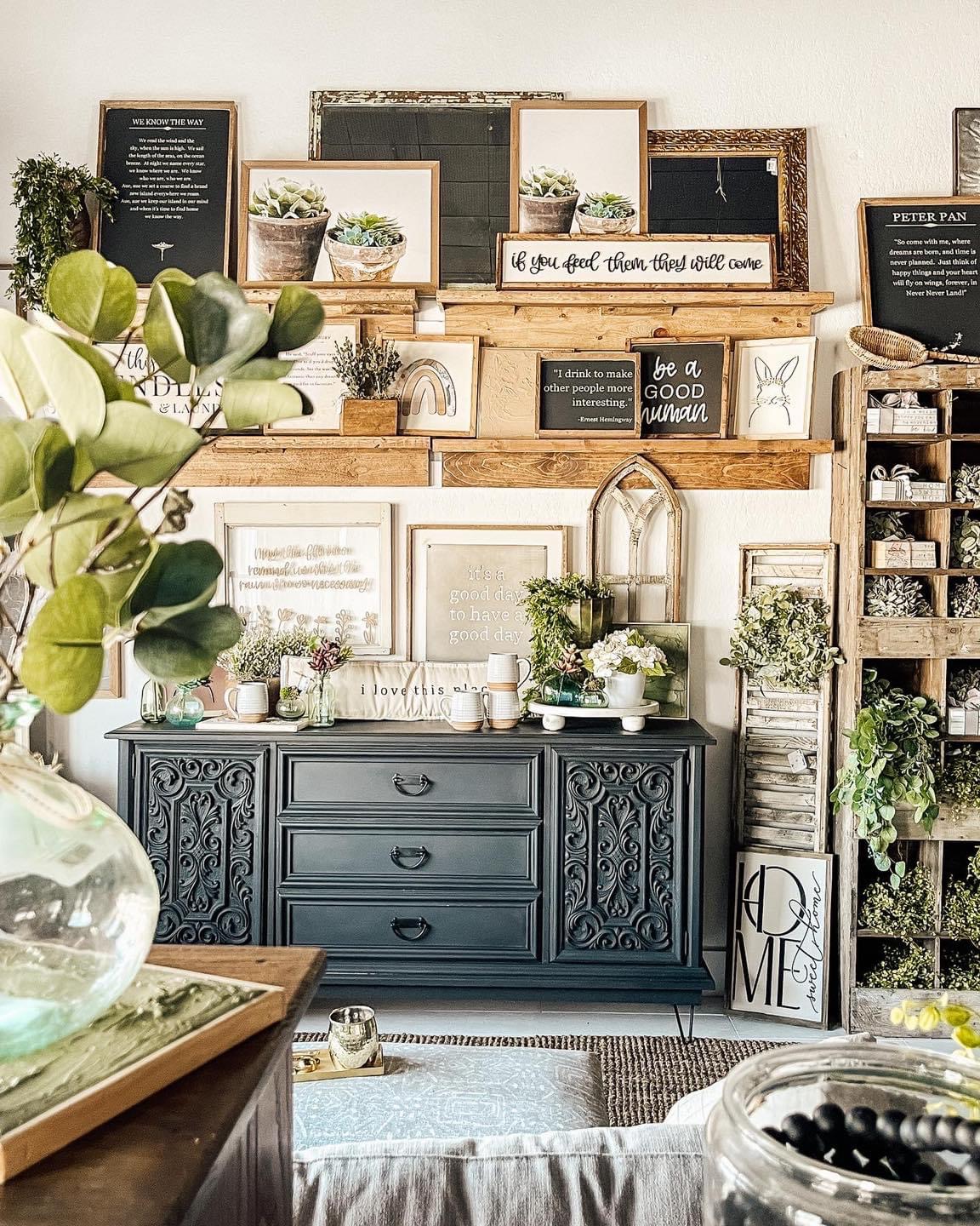 Decorative wall with framed pictures, shelves, plants, and a dark blue dresser with drawers, in a cozy interior setting.