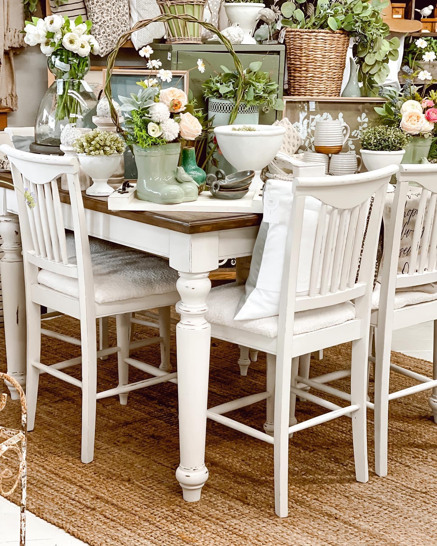Dining table with white chairs, decorated with flowers, vases, and bowls, in a cozy room with a woven rug.