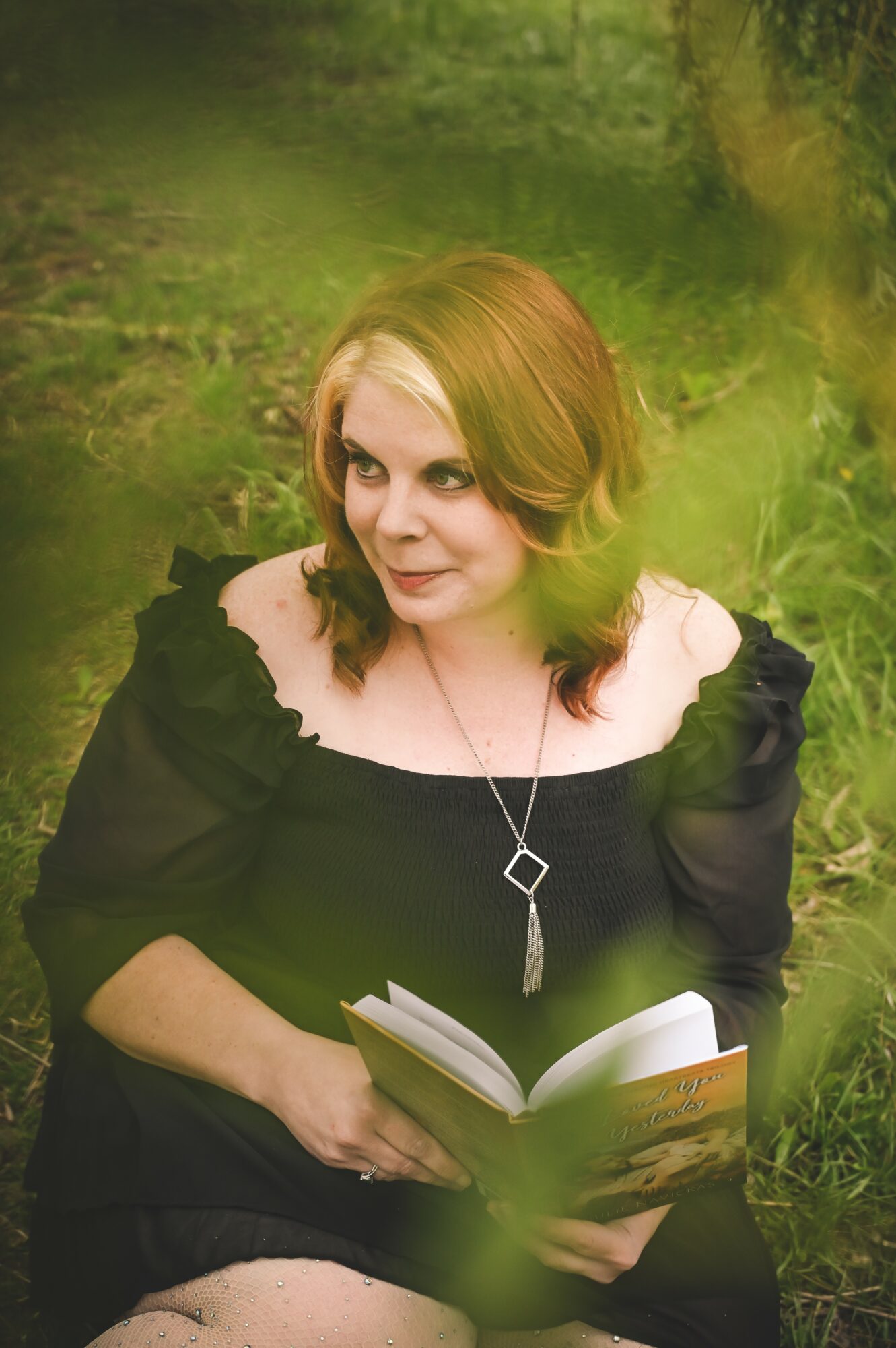 Woman with red hair in a black dress sitting outdoors, holding an open book, surrounded by greenery.
