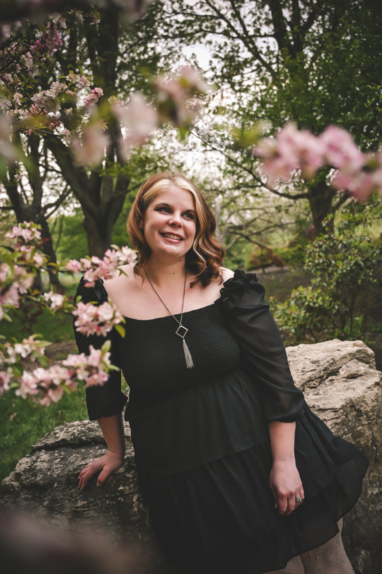 Smiling woman in black dress outdoors with trees and pink flowers, rocks in background.