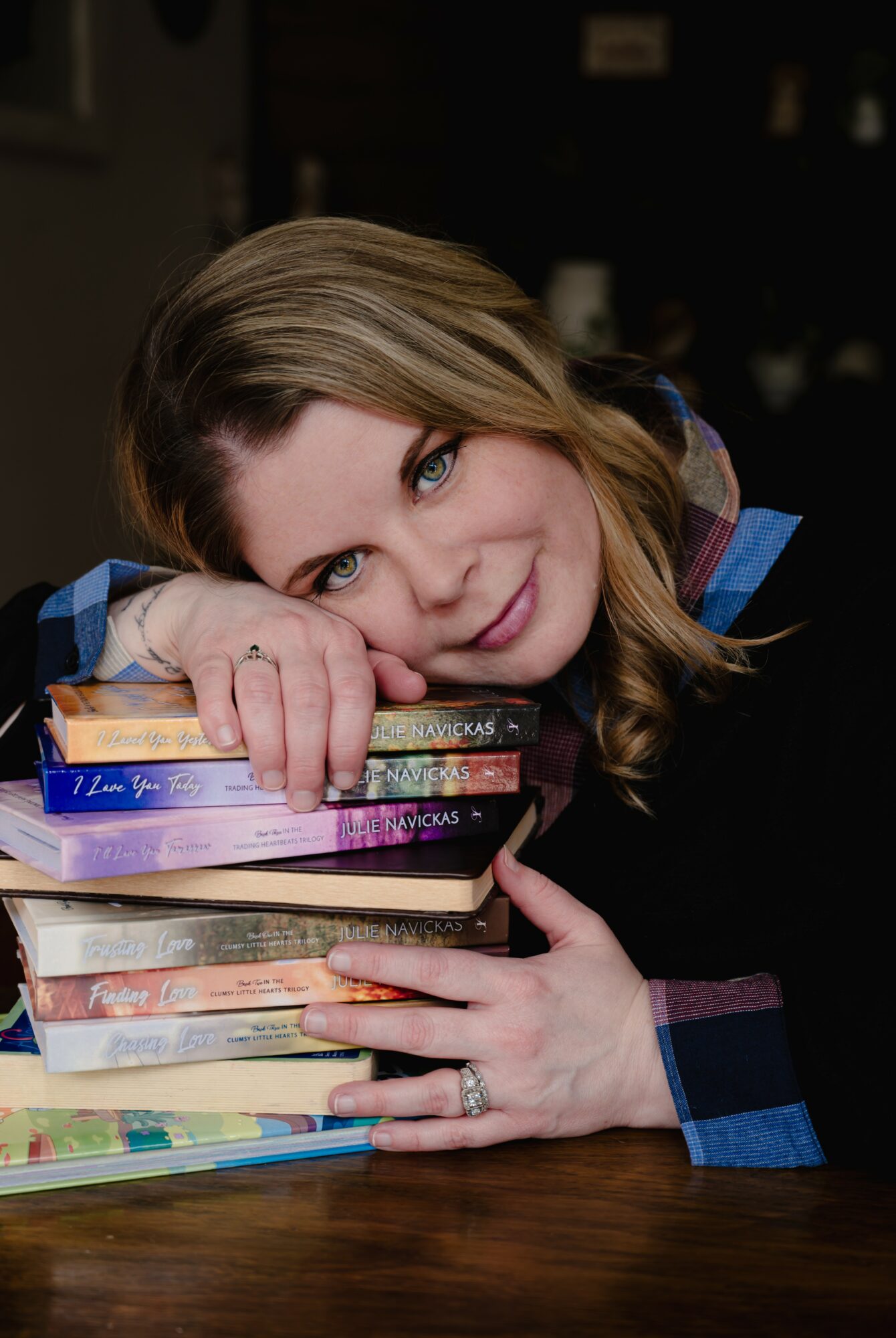 Woman resting her head on a stack of books, smiling, with dark background, long wavy hair, and colorful clothing.