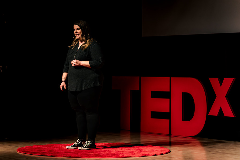 Woman standing on stage with TEDx sign behind her, speaking or presenting, on a red circular carpet, dark background.