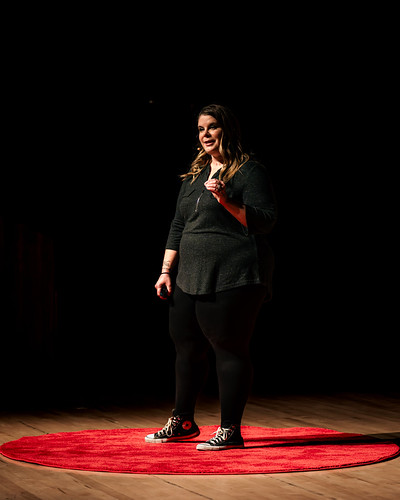 Woman standing on a red circular carpet on stage, smiling, with a dark background.