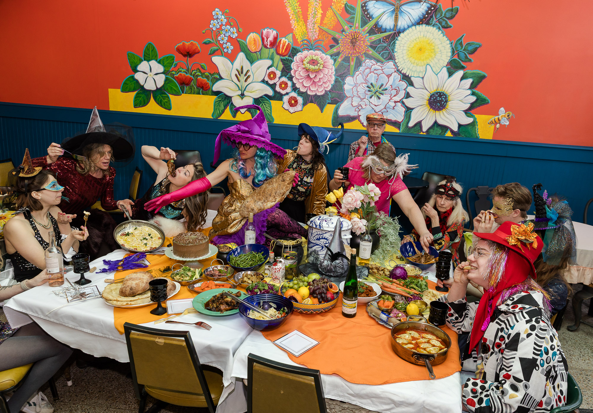 People in colorful costumes gather around a table with food and decorations, with a floral mural on the wall behind them.