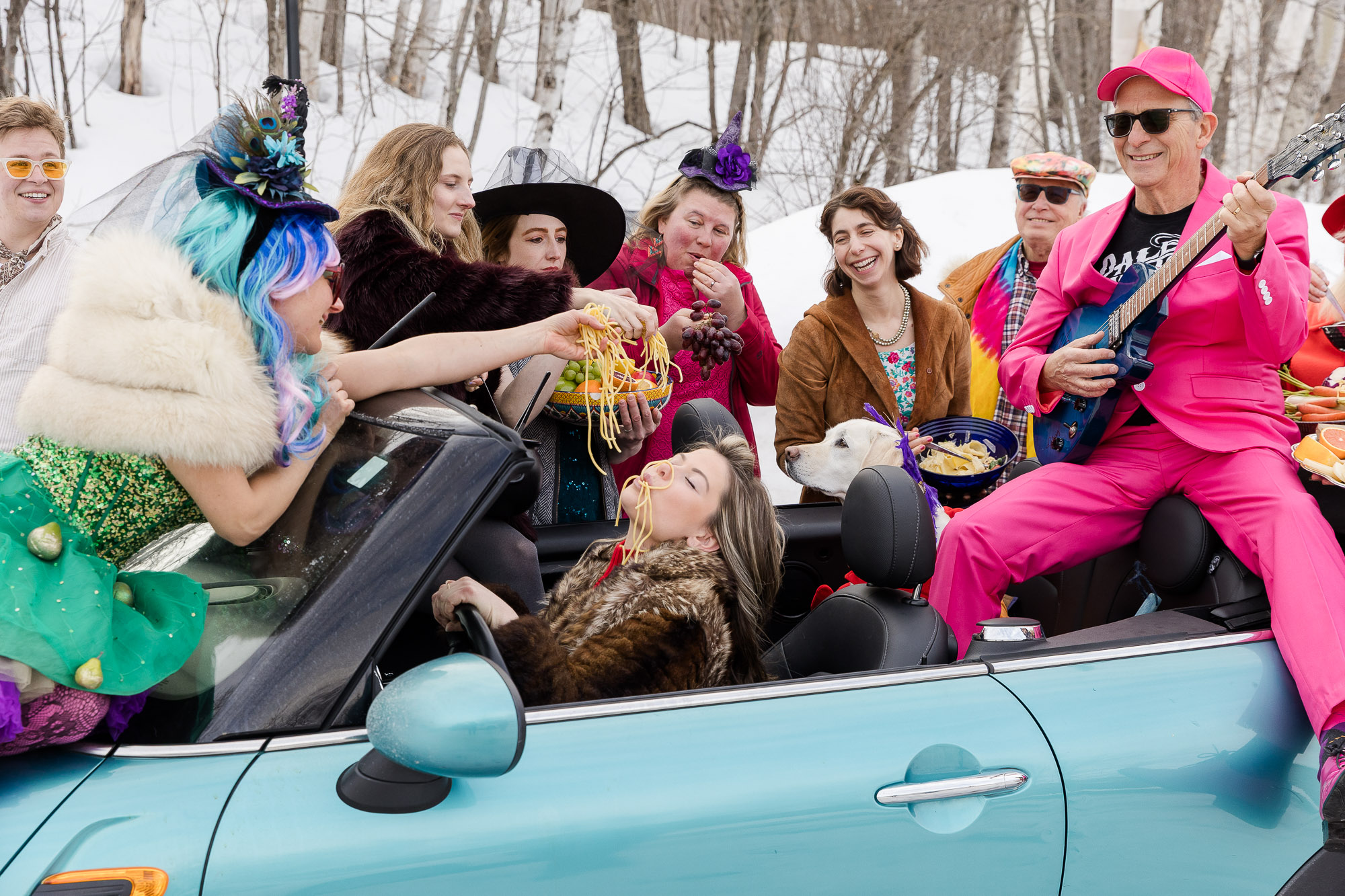 Group of people in colorful costumes riding in a convertible car outdoors in winter, some wearing hats and accessories.