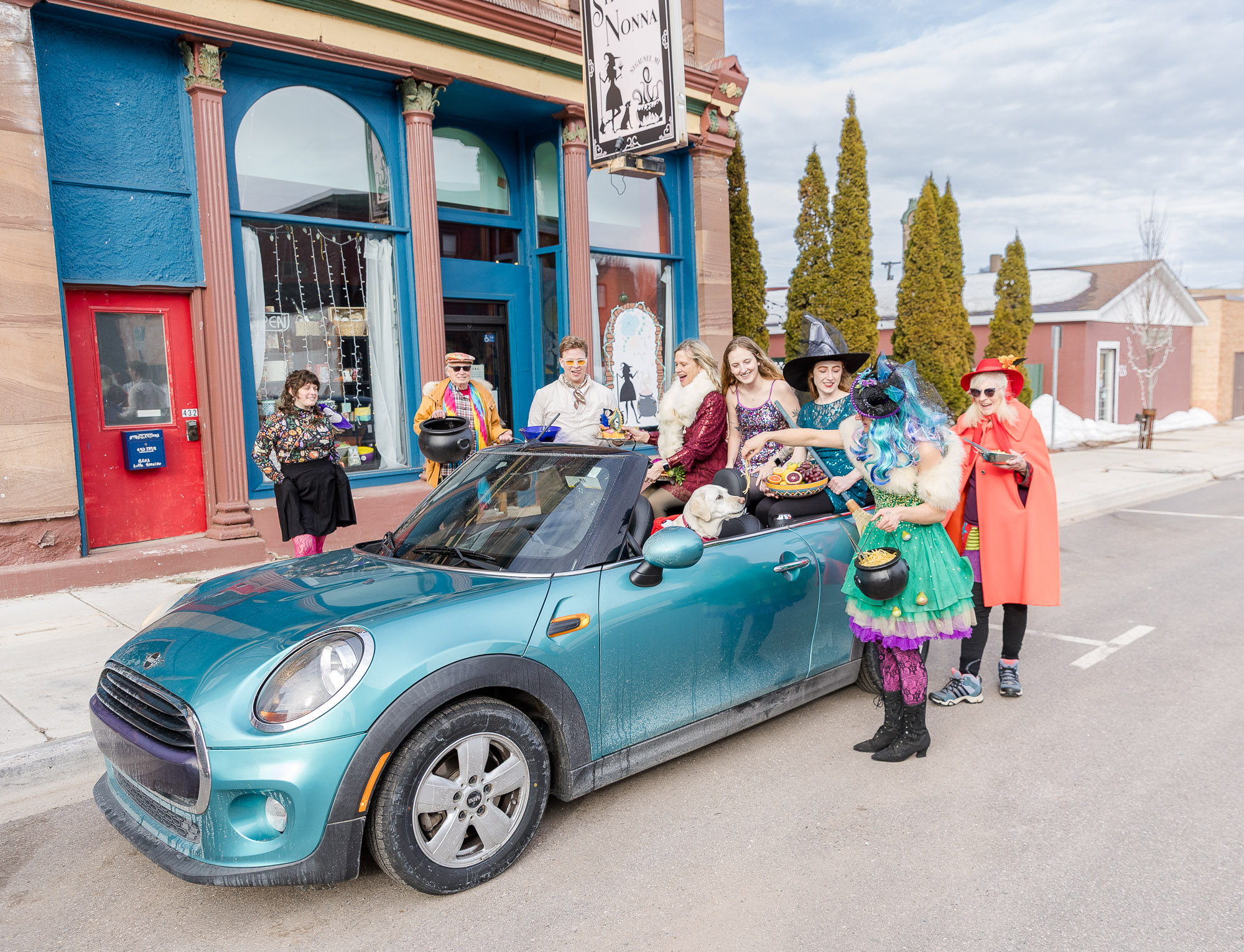 Group of people in colorful costumes gathered around a teal convertible car on a street with a colorful building and trees in the background.