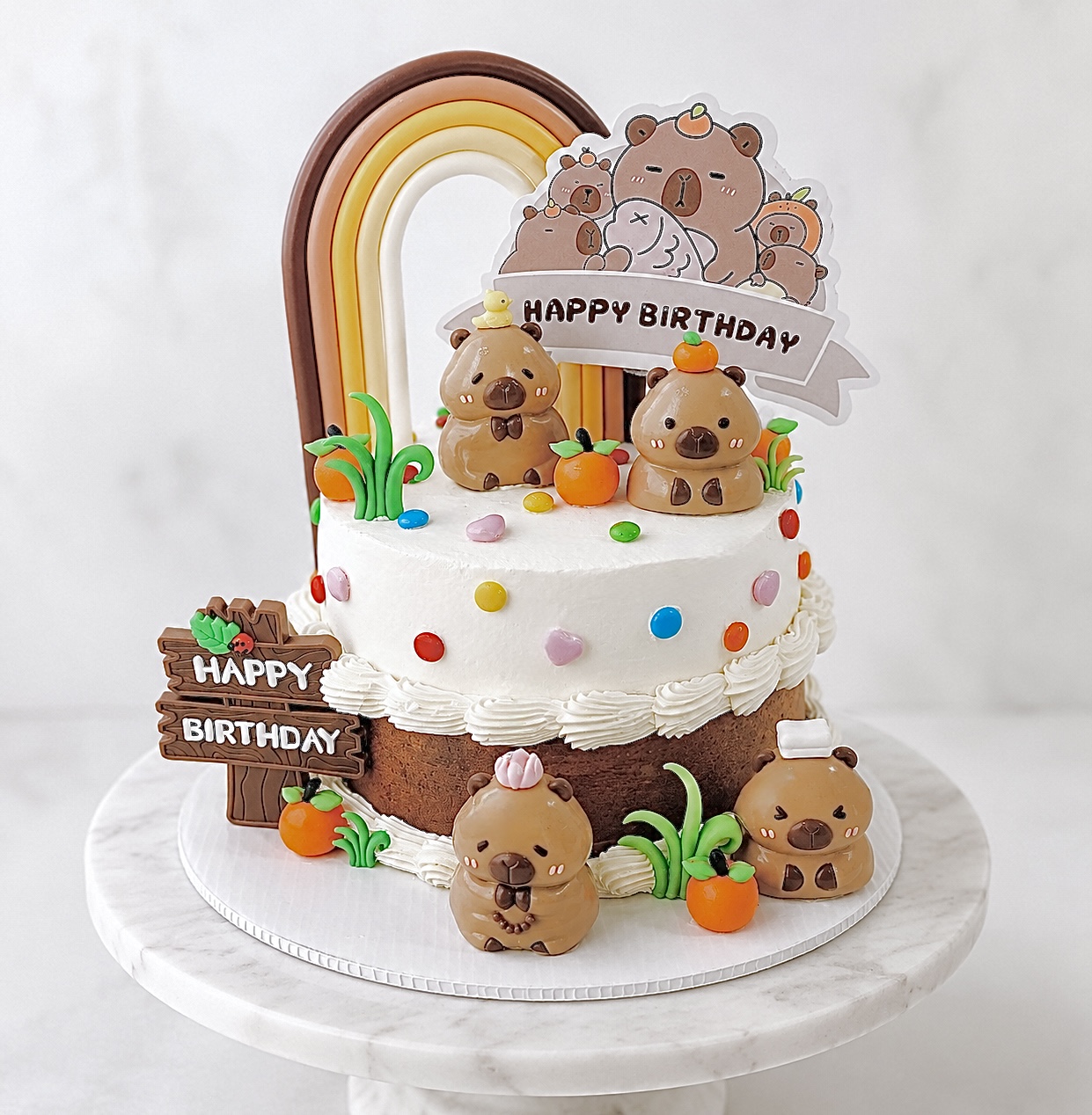 Decorated birthday cake with bear and rainbow toppers, colorful sprinkles, and small pumpkins, on a white marble surface.