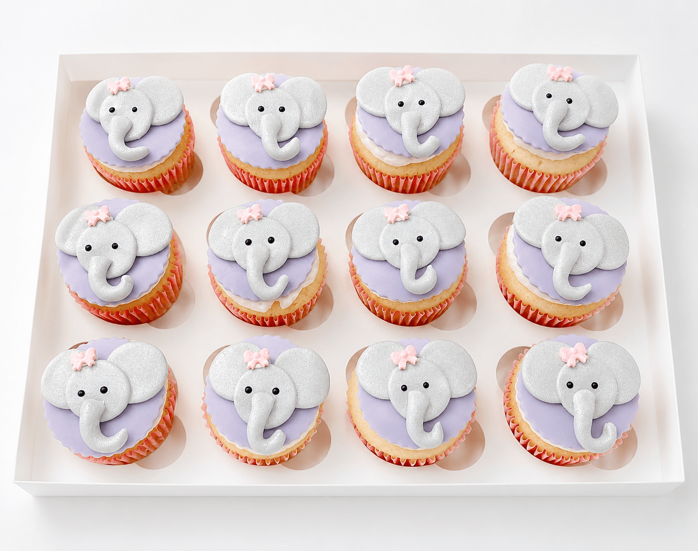 Twelve cupcakes with elephant decorations and pink bows, arranged in three rows on a white tray.