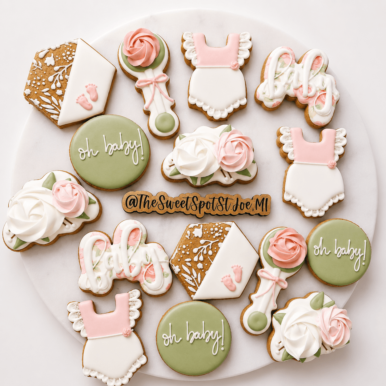 Assorted decorated cookies with pink, white, and green colors, including dresses, roses, and a cake with 'Oh baby!' written, on a white background.