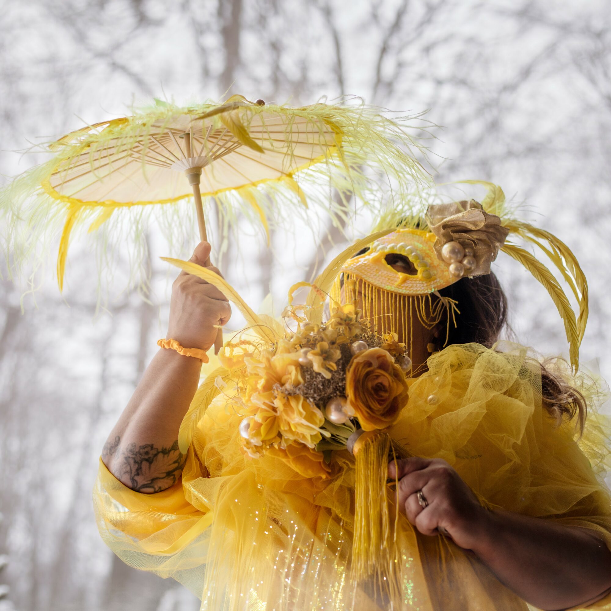 Person dressed in yellow with a floral headpiece holding a yellow parasol outdoors, face obscured by a decorative mask.