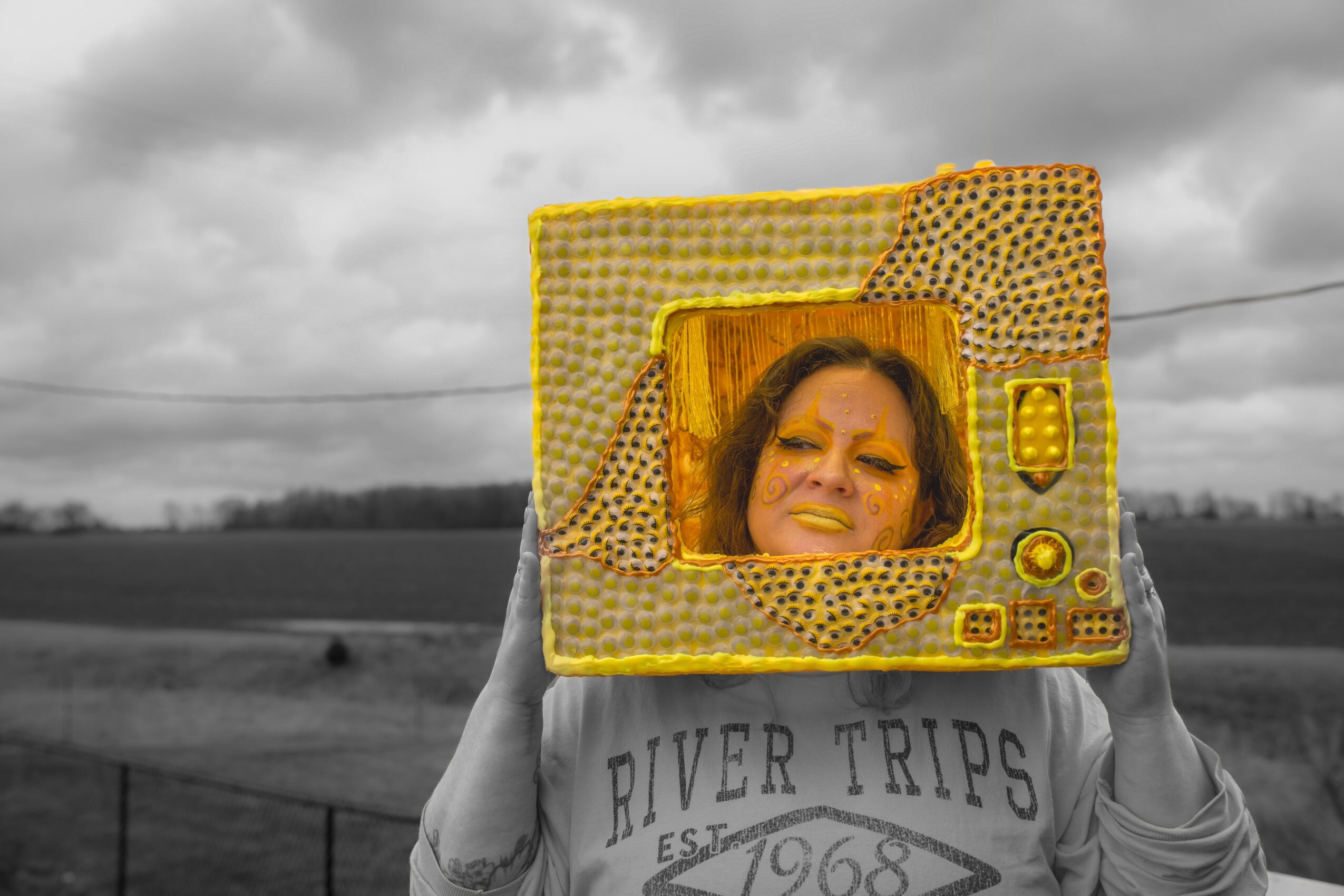 Person holding a yellow, vintage-style television with a woman's face on the screen, outdoors with cloudy sky and fields in background.