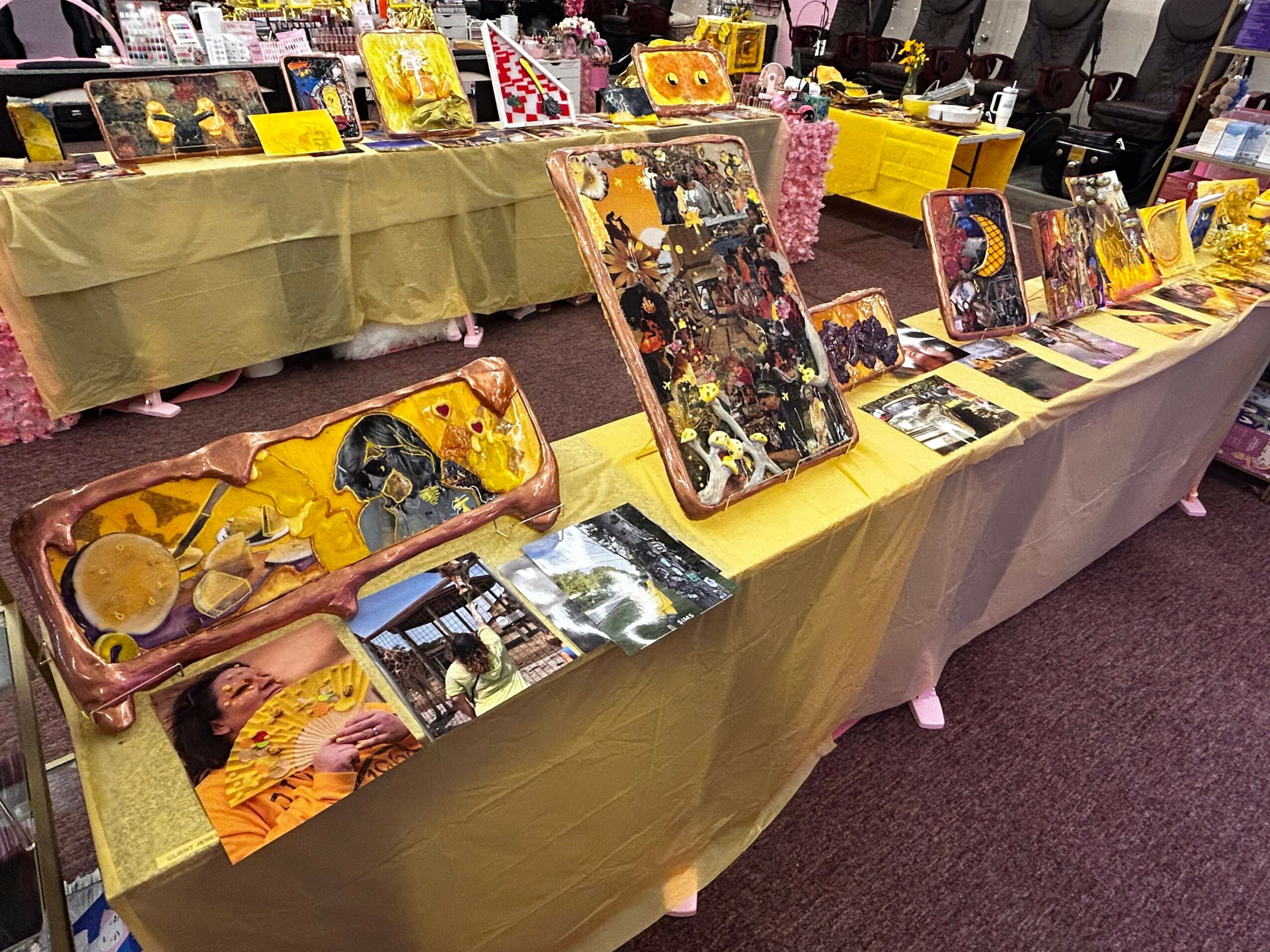 Tables covered with yellow cloth display colorful paintings and photographs at an indoor event.