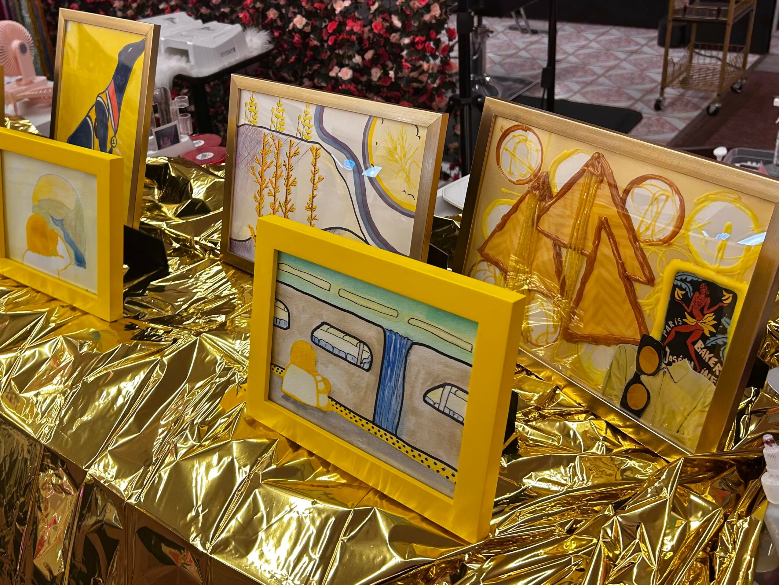 Display of colorful framed artwork on a table covered with gold foil, with various paintings and drawings visible.