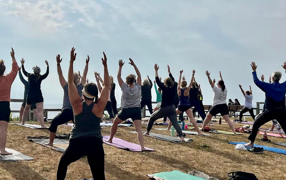 Group of people practicing yoga outdoors on mats, raising arms in a pose, with a cloudy sky in the background.
