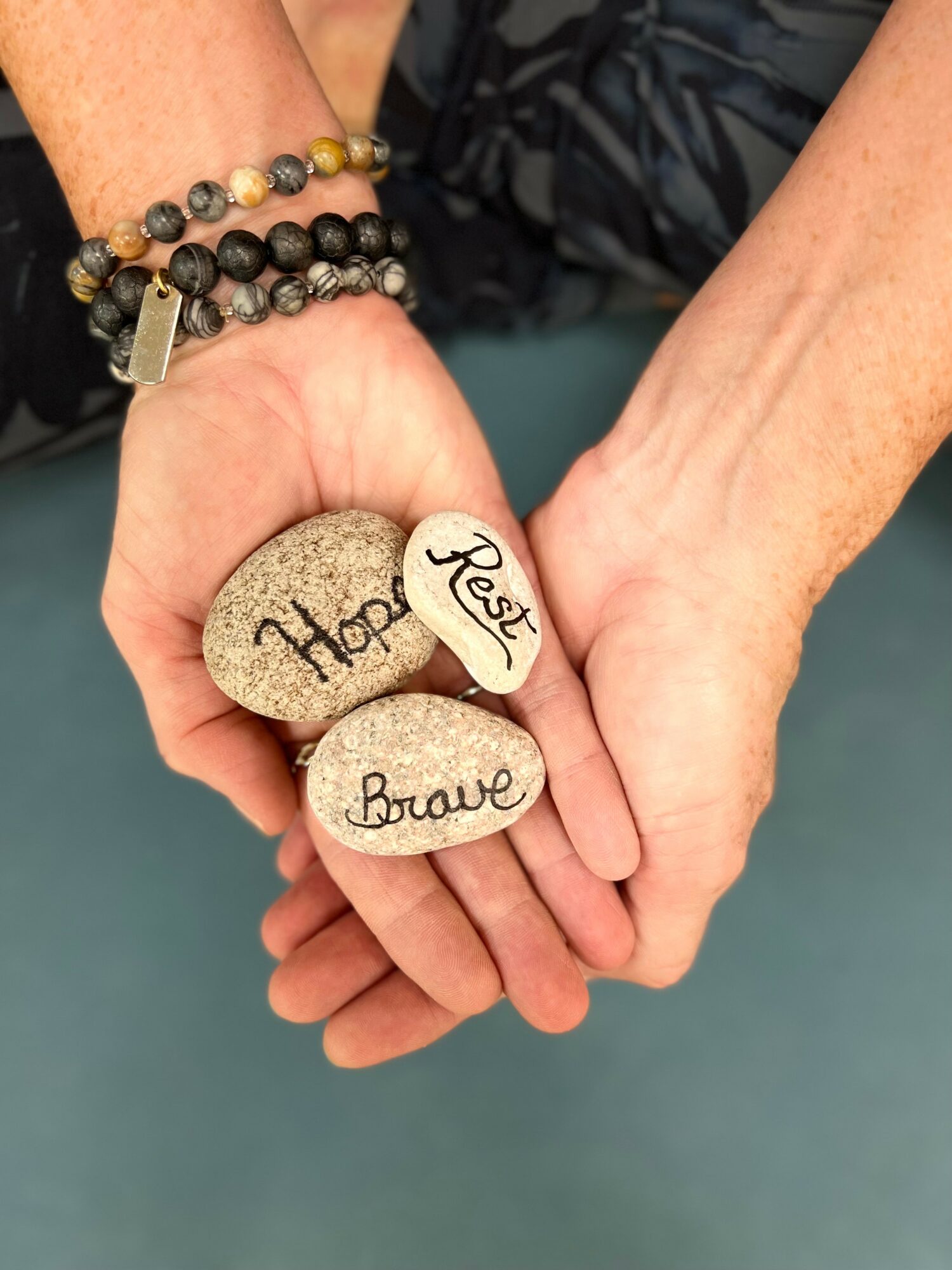Two hands holding three stones with words: Hope, Rest, Brave, in a gentle clasp.
