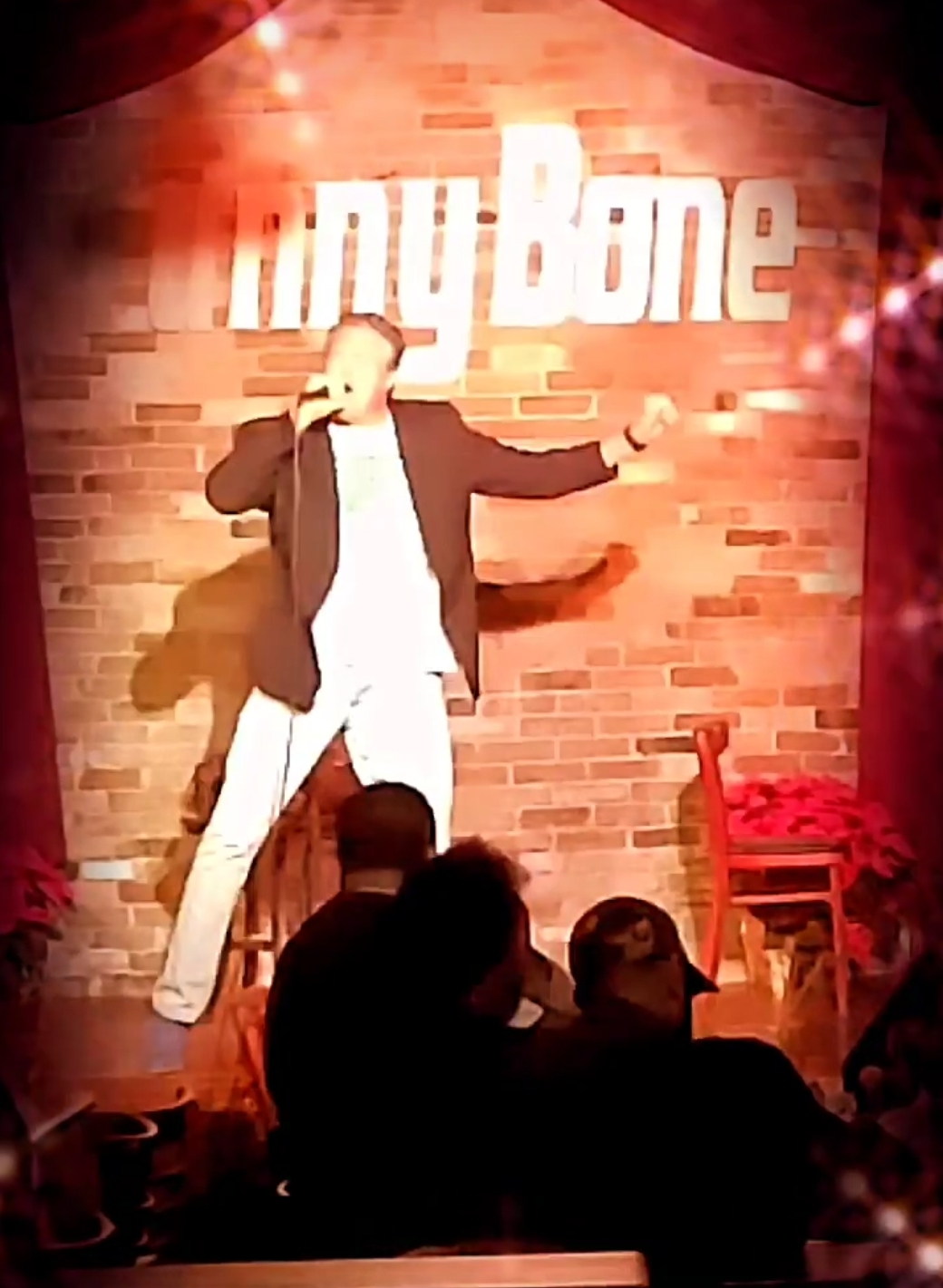 Performer on stage with a brick wall background and a red chair, holding a microphone, with audience in foreground.