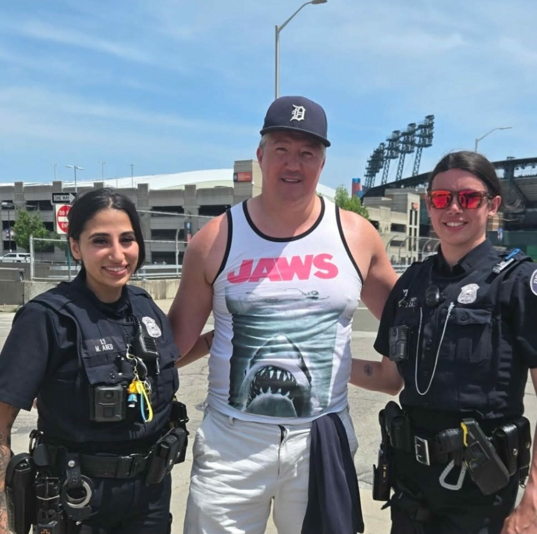 Two police officers and a man in a tank top with a shark graphic, standing outdoors with a stadium in background.