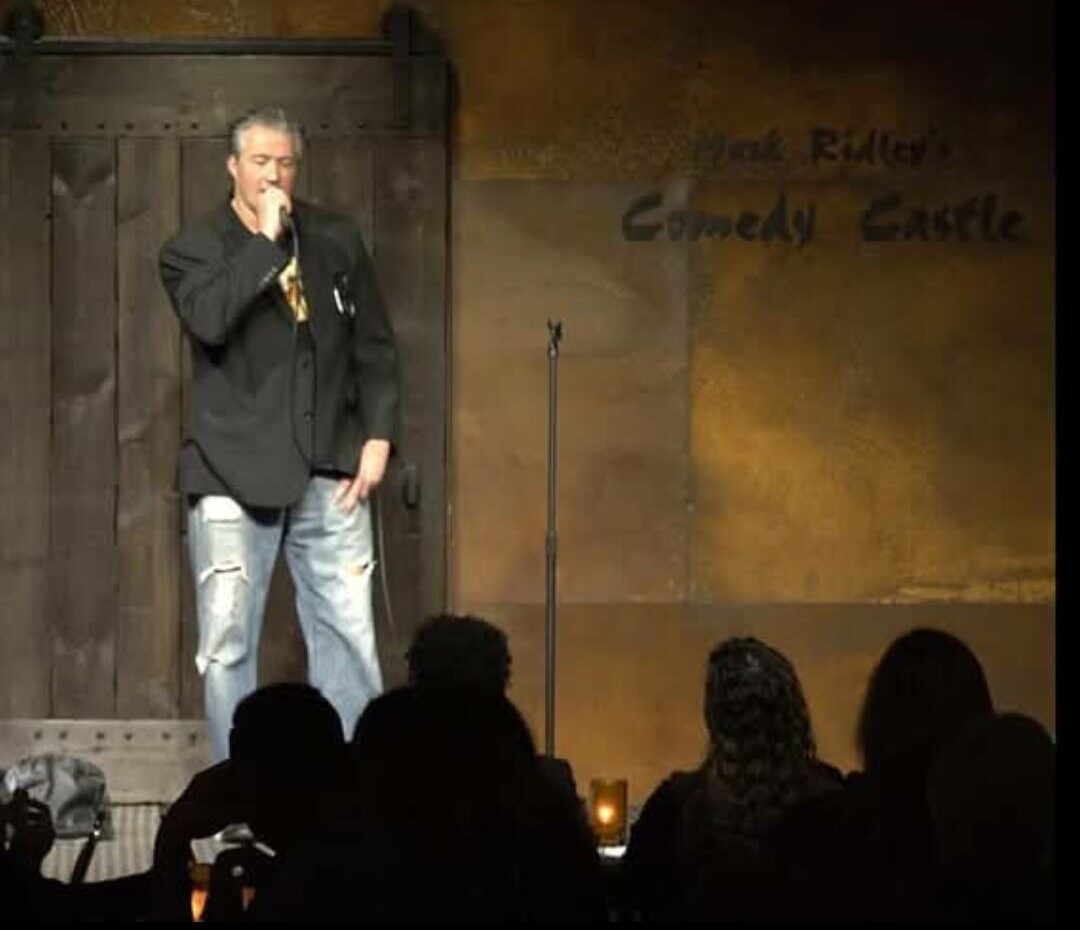 Person standing on stage speaking into microphone, audience silhouettes visible, background with text, dim lighting.