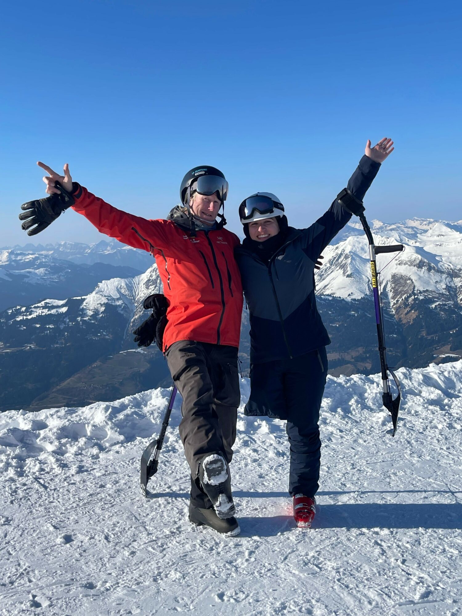 Two people in winter clothing standing on snow-covered ground with mountains in the background, one making a peace sign, the other raising an arm.