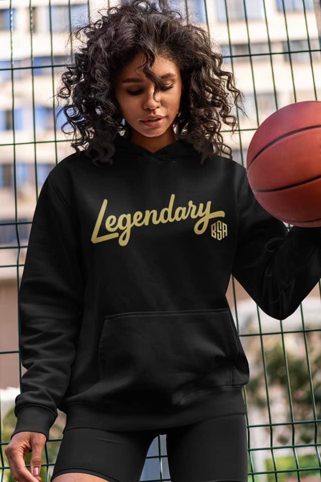 Young woman with curly hair holding a basketball on a court, wearing a black hoodie with 'Legendary' text.