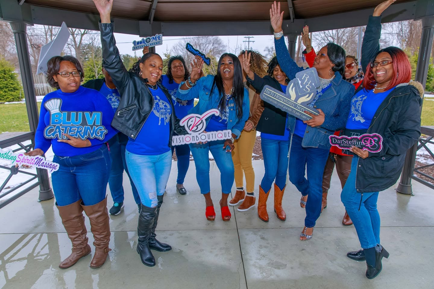 Group of women celebrating indoors, some raising hands, holding signs, and smiling, with large windows and outdoor scenery in background.