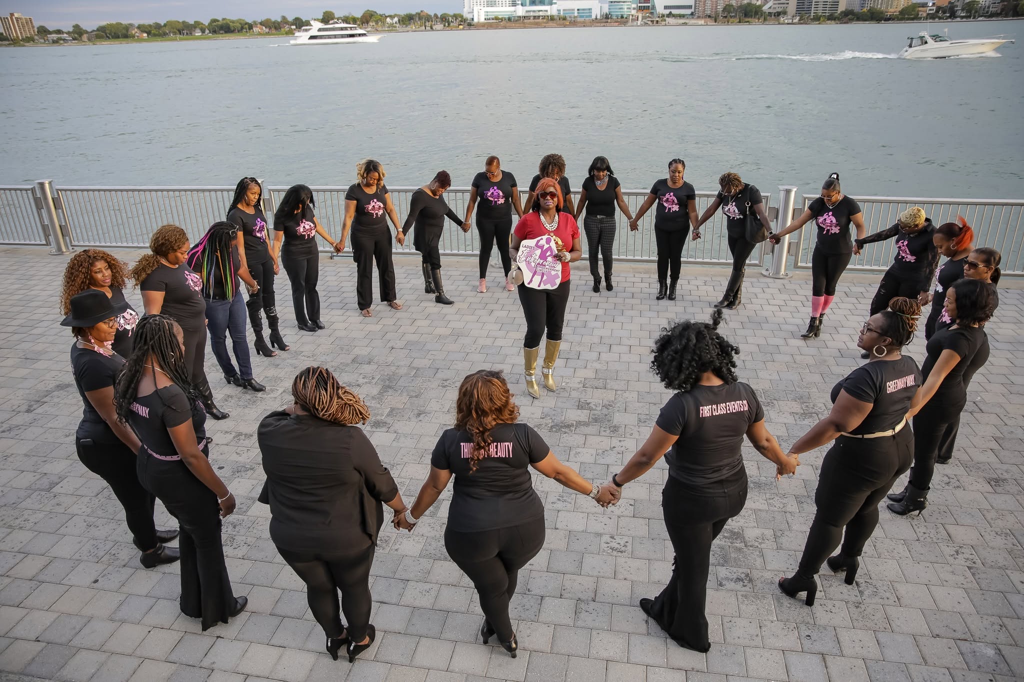 Group of women holding hands in a circle outdoors near water, some wearing matching black shirts, sunny day.