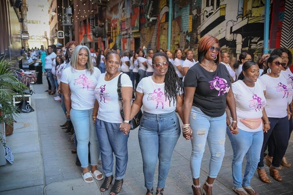 Group of women walking together on a city street, holding hands, with colorful graffiti in the background.