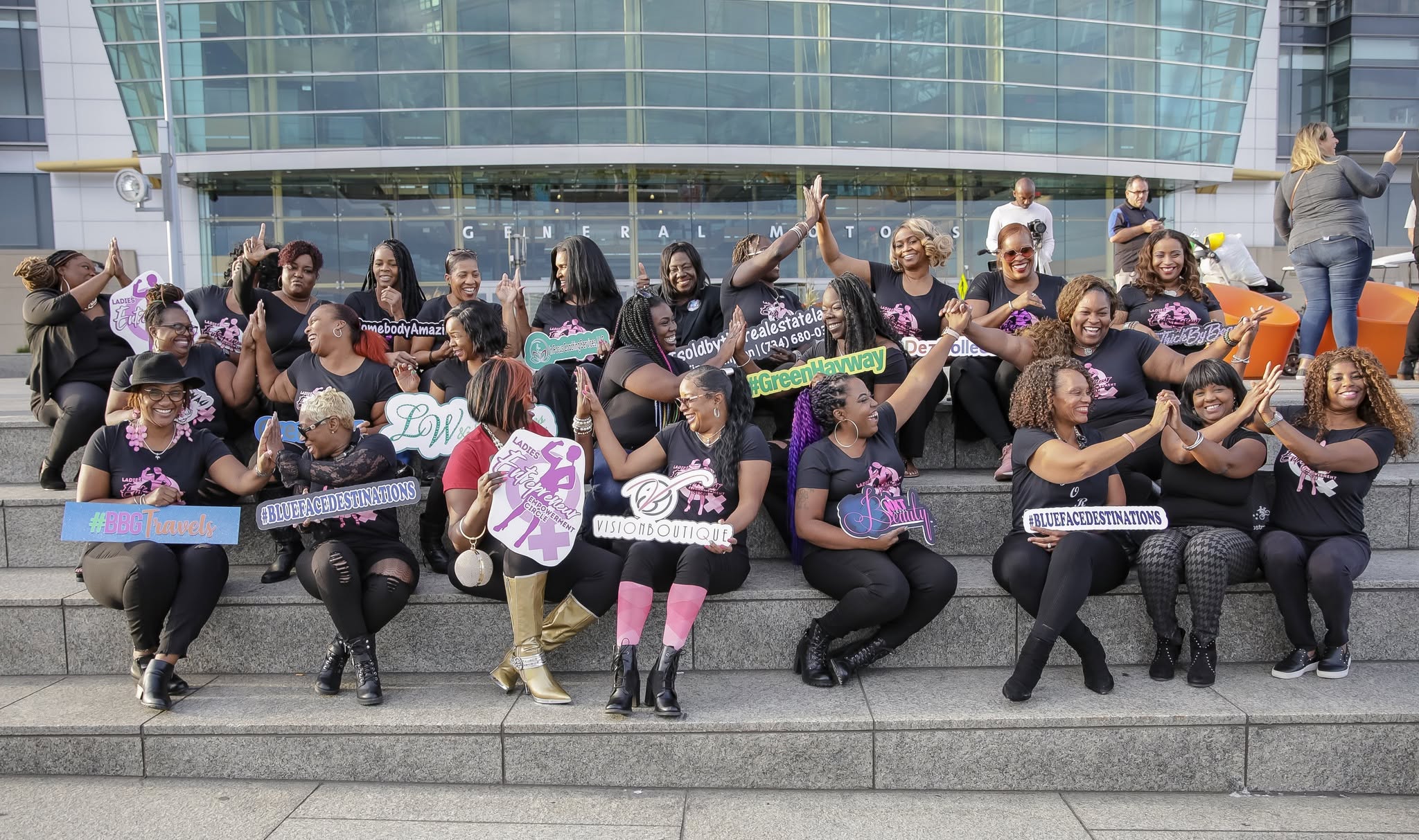 Group of women sitting on steps holding colorful signs and banners, smiling and waving, in front of a modern glass building.