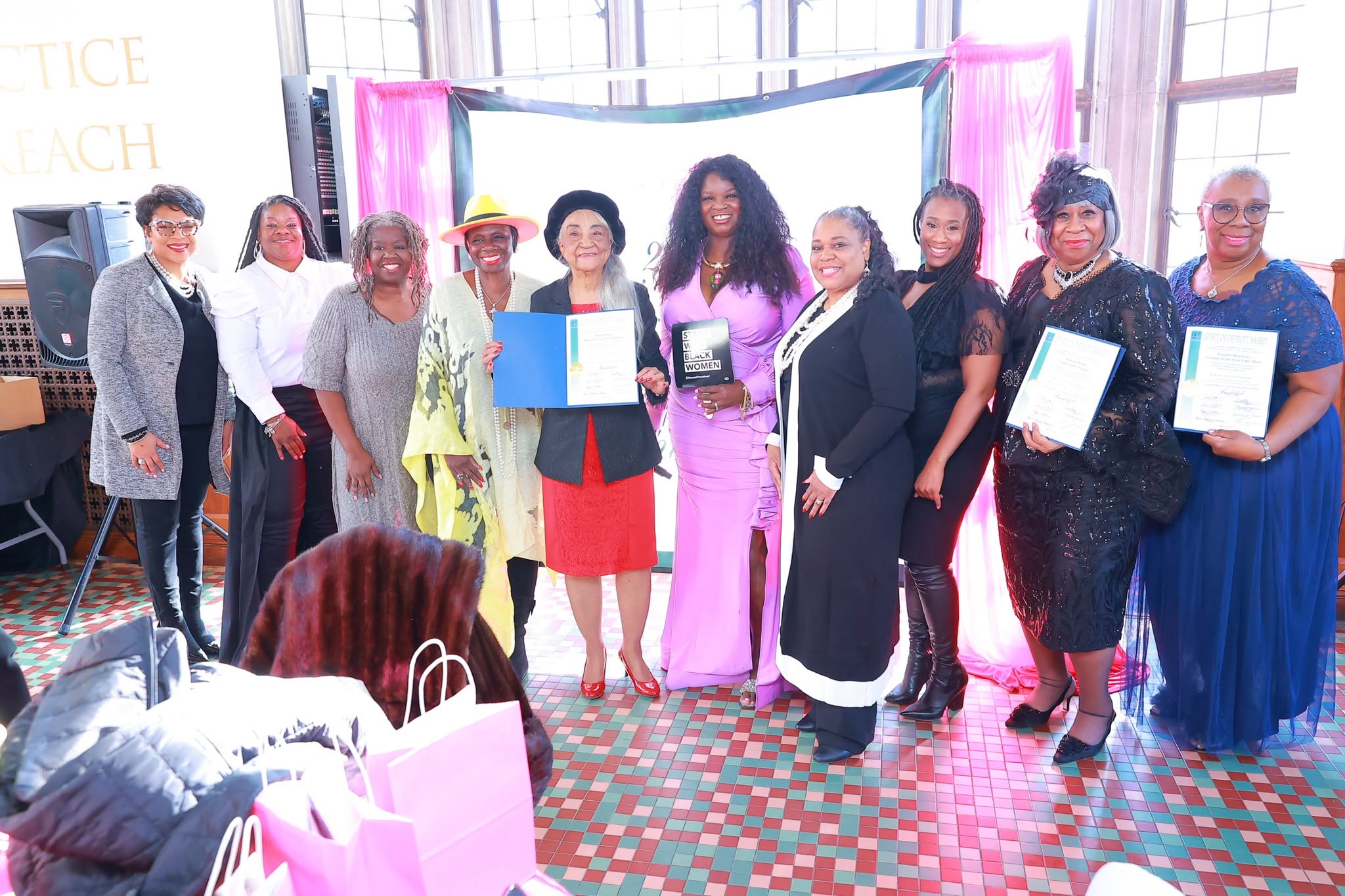 Group of ten women standing indoors, some holding certificates, in front of pink curtains and a window, smiling.