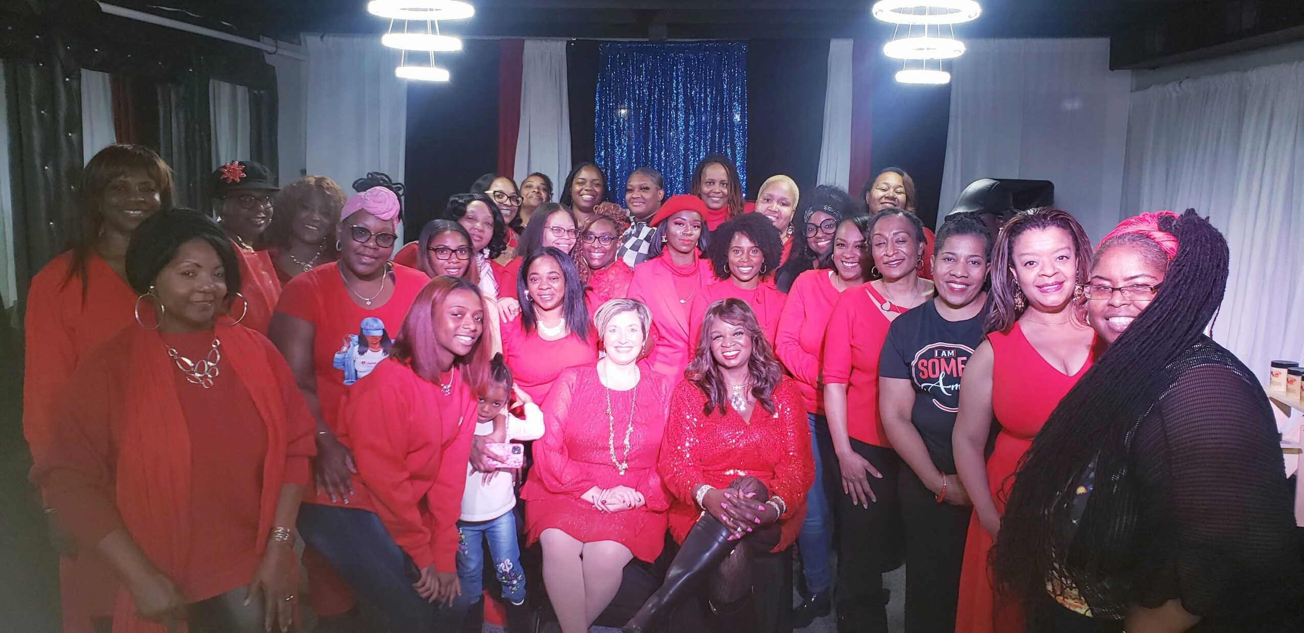 Group of people gathered indoors, many wearing red, smiling, with bright lights and curtains in the background.