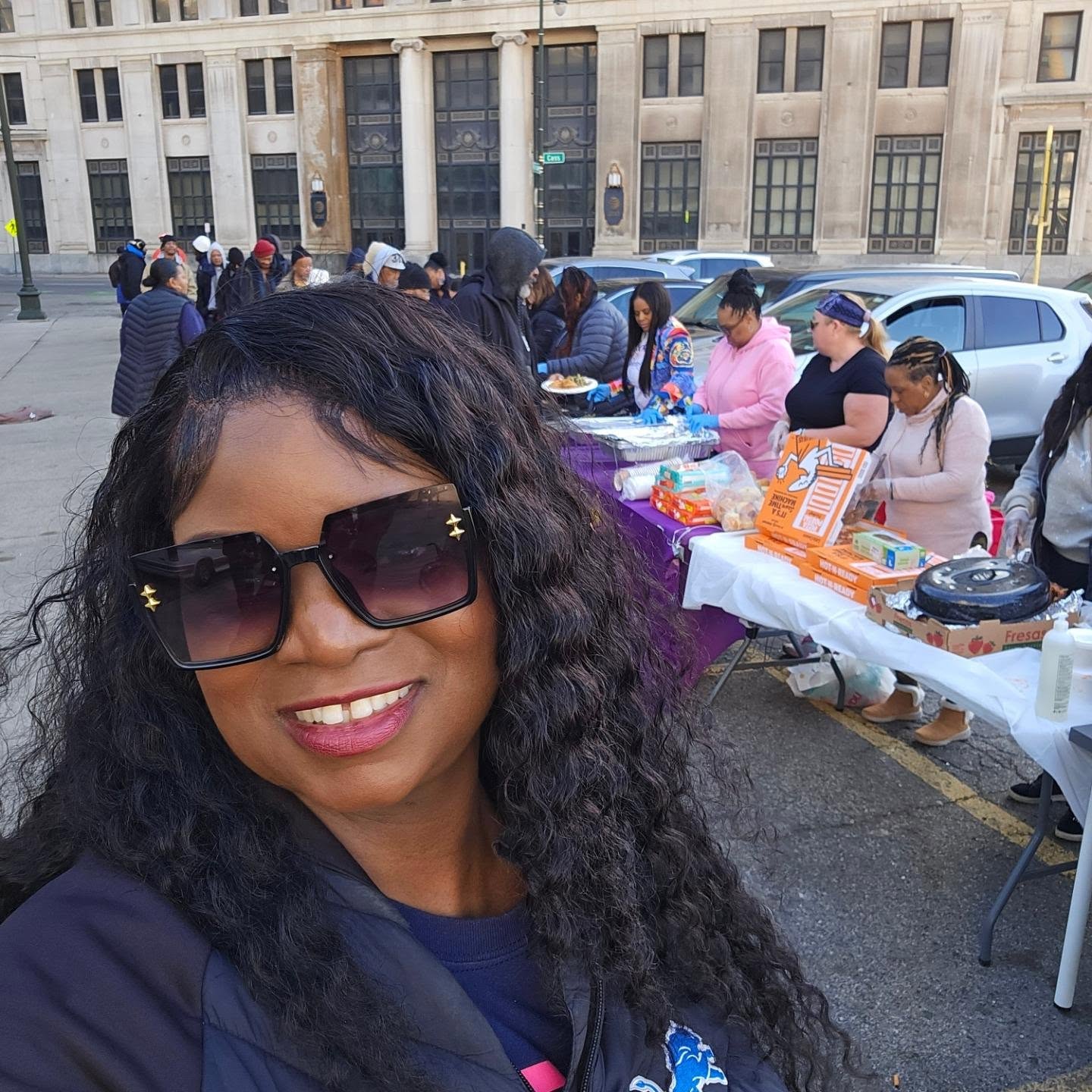 Woman with curly hair and sunglasses smiling in front of outdoor market with people and tables.