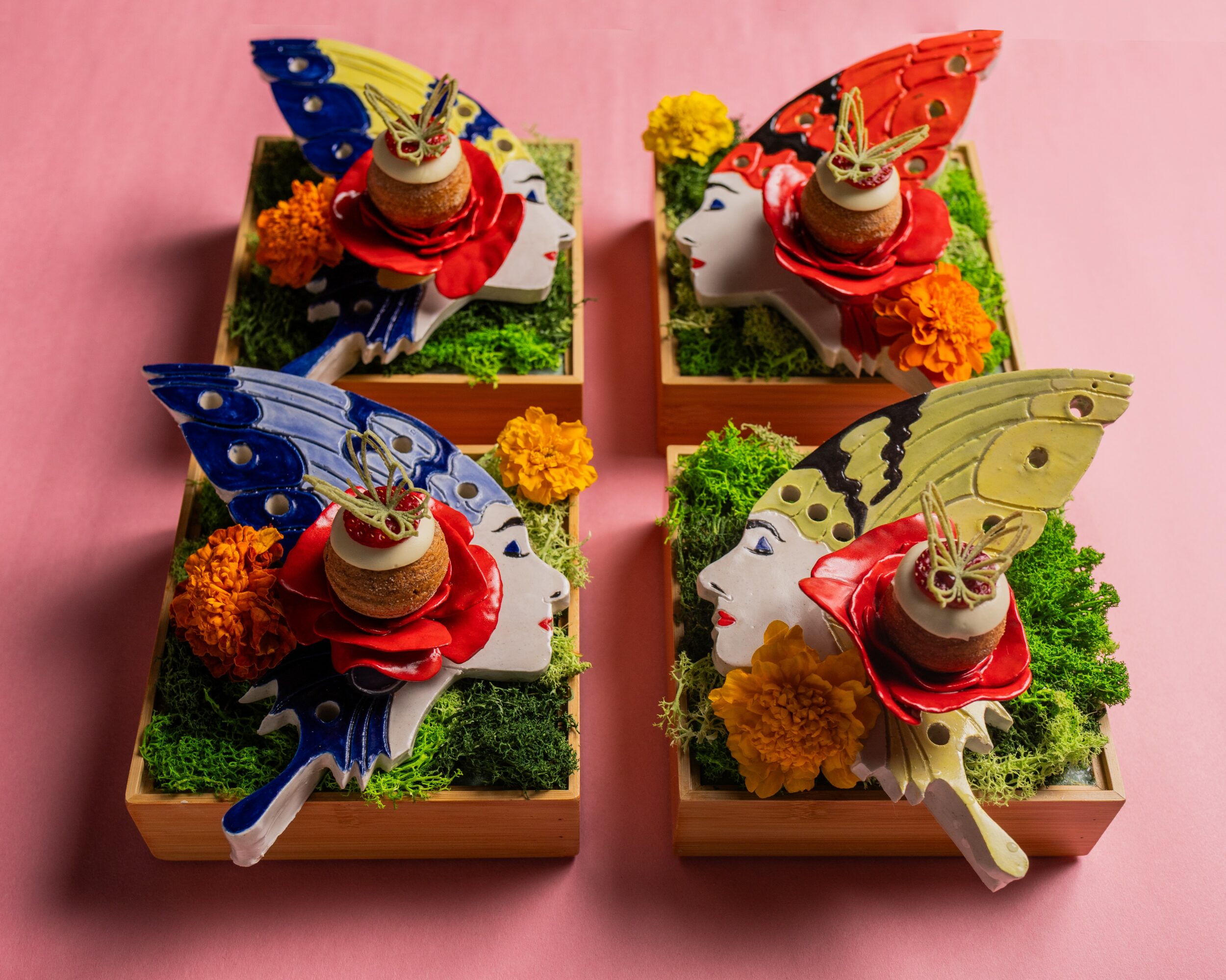 Four decorated boxes with butterfly and face designs, flowers, and green grass-like material, arranged on a pink background.