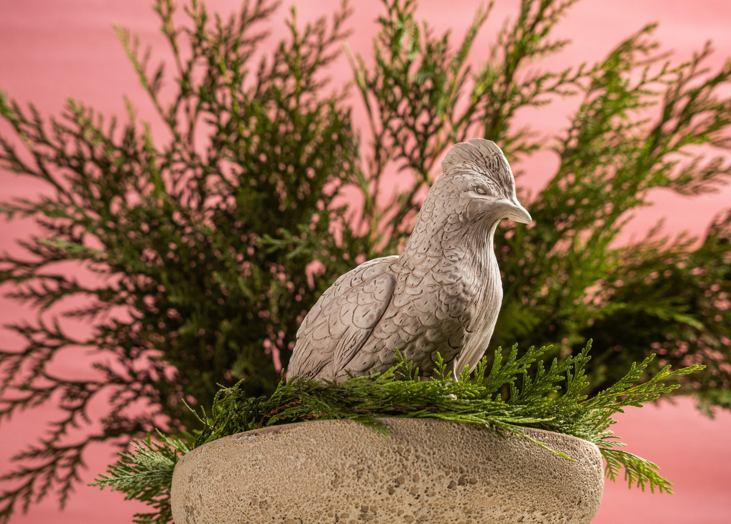 A bird sculpture on a stone pedestal with greenery, pink sky background, and trees behind.