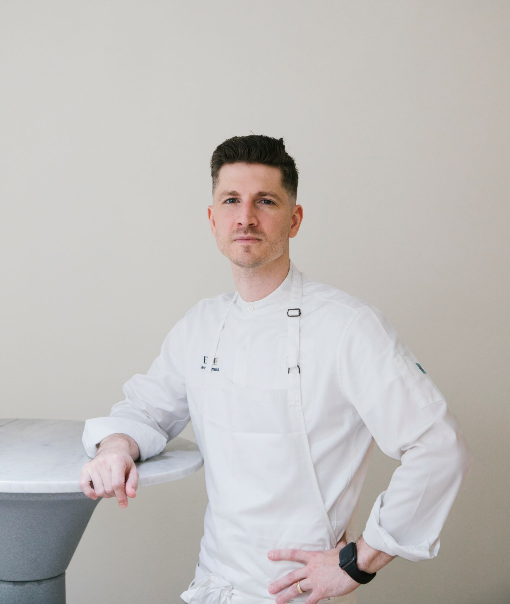 Male chef in white uniform standing with hand on hip, against plain light background.