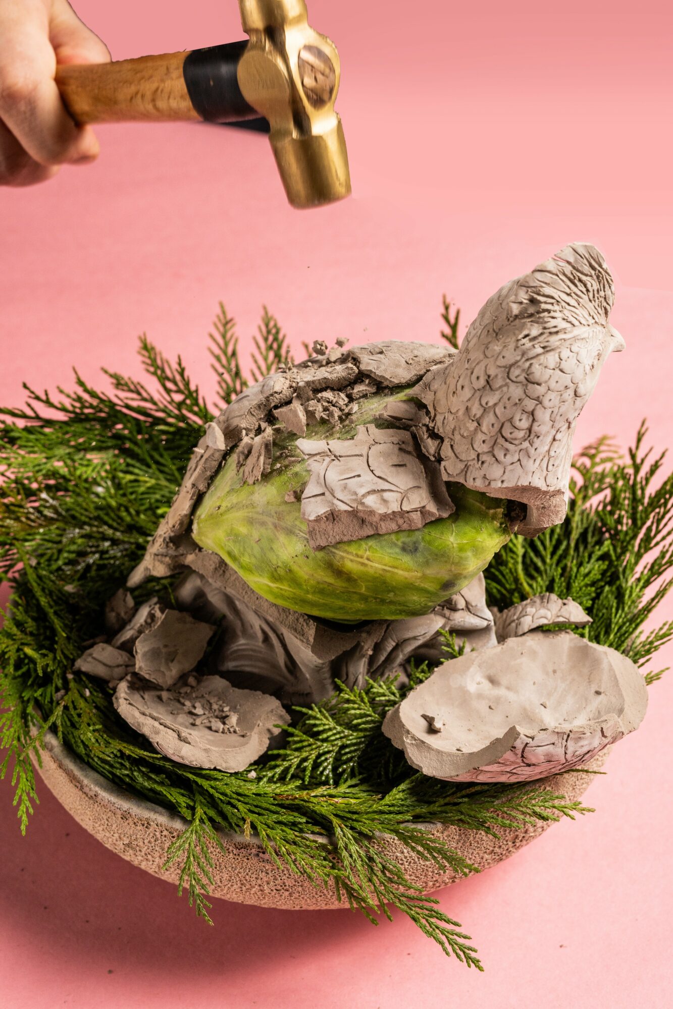 A hammer is about to hit a stone sculpture of a bird, surrounded by greenery on a pink background.