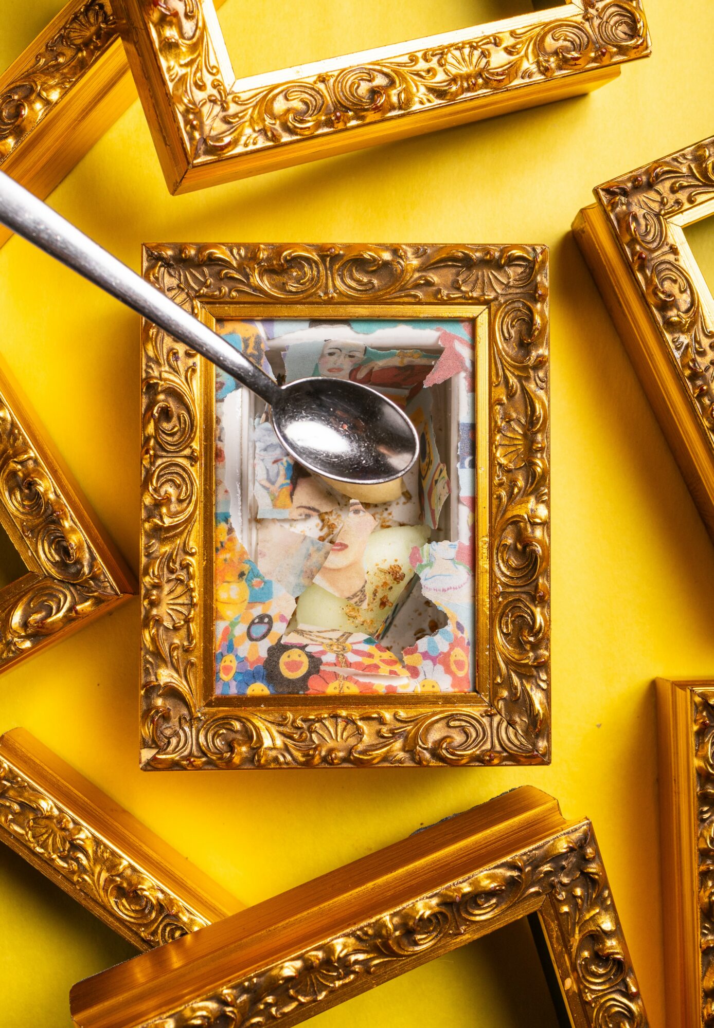 A spoon rests inside a picture frame with a colorful collage, surrounded by other ornate frames on a yellow background.
