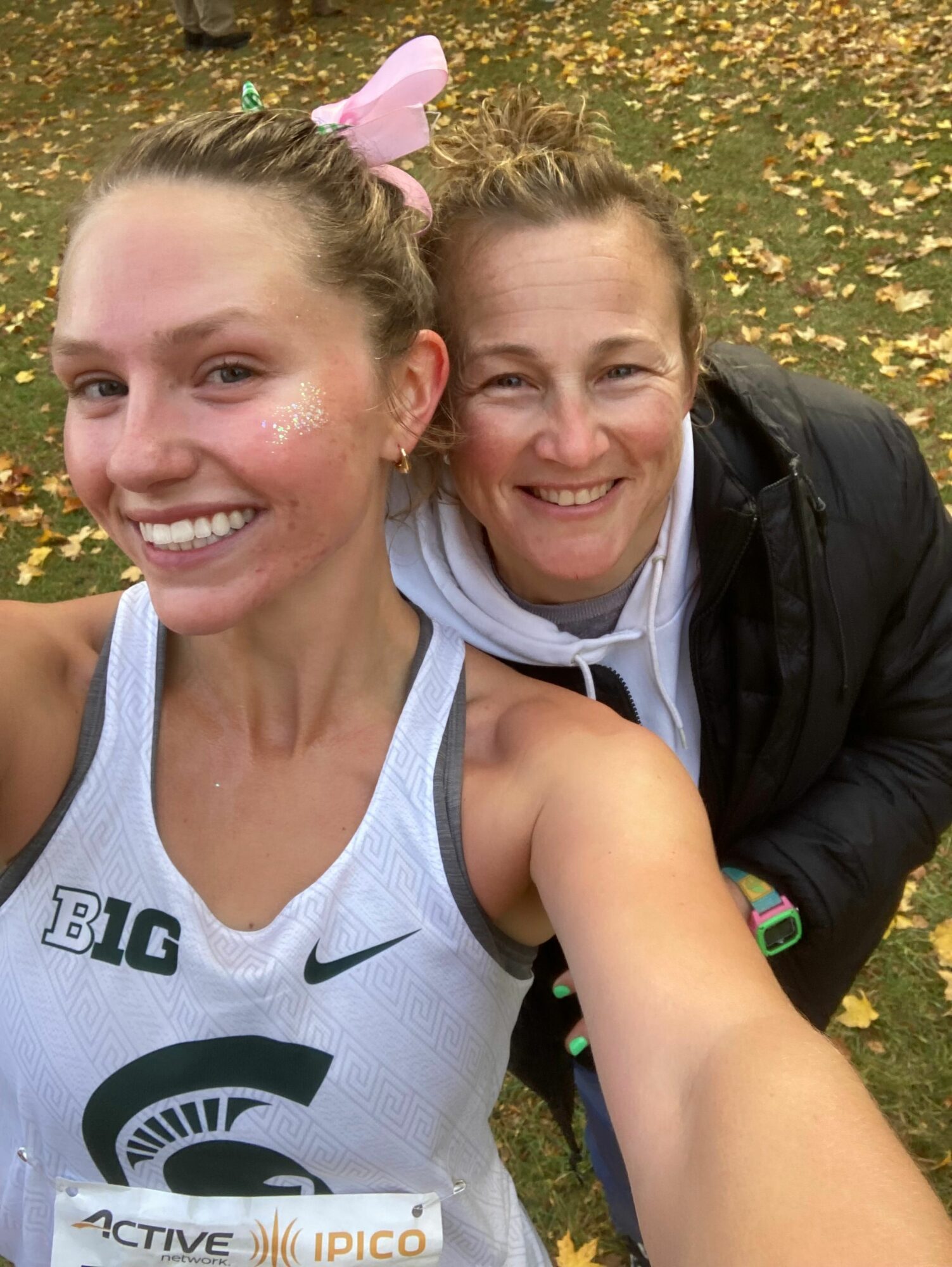 Two smiling women taking a selfie outdoors, one wearing a white sports top, the other in a black jacket, with trees and yellow flowers in the background.