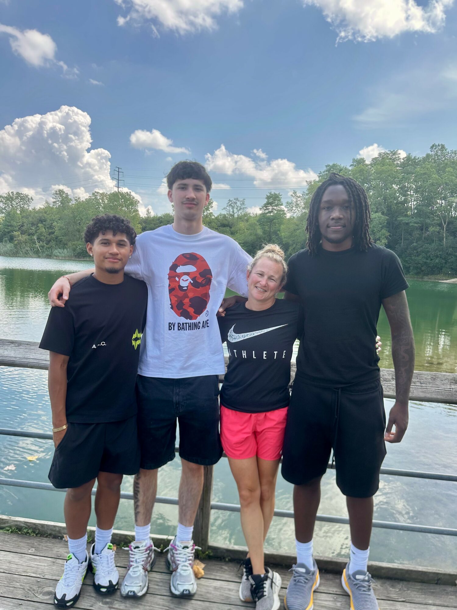 Four people standing outdoors by a lake with trees and clouds in the background, smiling, with arms around each other.